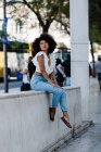 Attractive ethnic woman in jeans and tank top relaxing on stone railing against urban background — Stock Photo