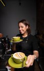 Усміхнена бариста дає зелені чашки кави з латте мистецтвом і дивиться на камеру в кафе — стокове фото