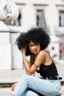 Portrait of ethnic woman in jeans and tank top relaxing and sunbathing against urban background — Stock Photo