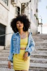 Donna afroamericana in abito giallo e giacca di jeans in piedi sulle scale e guardando la fotocamera su sfondo urbano — Foto stock