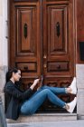Вид збоку красивої жінки, використовуючи цифровий планшет, розслабляючись на порозі старих дерев'яних дверей — стокове фото
