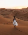 Vista posteriore delle donne scalze in abito bianco estivo che camminano su dune sabbiose di deserto infinito al tramonto, Marocco — Foto stock