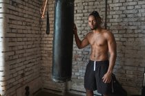 Уверенный чернокожий боксер в спортзале позирует с боксерской сумкой — стоковое фото
