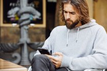 Jeune homme beau barbu en pull décontracté assis au café concentré sur le téléphone mobile — Photo de stock