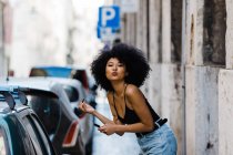 Portrait of young ethnic woman standing near car window and doing makeup while kissing at camera on urban background — Stock Photo