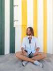 Allegro uomo barbuto in abito casual tenendo smartphone mentre seduto sulla sabbia contro parete a strisce sul resort — Foto stock