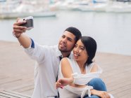 Jovem casal sentado e se divertindo tirando uma foto selfie no banco de madeira no litoral arenoso e beijando — Fotografia de Stock