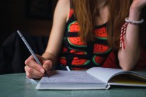 Femme de culture méconnaissable assis à la table dans une chambre confortable et l'écriture dans un carnet — Photo de stock