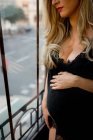 Невизначена жінка з світлим волоссям торкається вагітної животики і спирається на віконне скло вдома — стокове фото