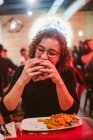 Mulher nova com fome mordendo hambúrguer saboroso enquanto sentado à mesa em café brilhantemente iluminado — Fotografia de Stock