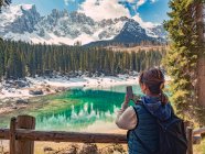 Back view of young woman in down vest shooting on phone majestic landscape of turquoise water surrounded by rocky mountains and forest in cloudy day — Stock Photo