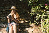 Mulher viajando em vestido e chapéu usando câmera enquanto sentado em degraus de pedra na rua ensolarada — Fotografia de Stock