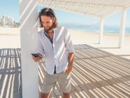 Handsome bearded man using smartphone while leaning on pillar of white gazebo on sandy beach — Stock Photo