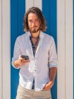 Glücklicher bärtiger Kerl mit Smartphone, während er an einem sonnigen Tag im Resort gegen eine gestreifte Wand steht — Stockfoto