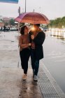 Веселий молодий чоловік і жінка з парасолькою, що приймає і дивиться один на одного, стоячи на мокрій міській вулиці в дощовий день — стокове фото
