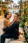 Вид збоку молодого чоловіка і жінки, що обіймає і дивиться на камеру, сидячи на лавці під час романтичного побачення на міській вулиці — стокове фото