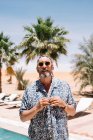 Bearded senior male in sunglasses buttoning shirt while standing on poolside on sunny day on resort — Stock Photo