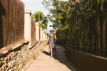 Glückliche Frau mit Strohhut und Sonnenbrille spaziert bei Sonnenschein durch die enge Straße — Stockfoto