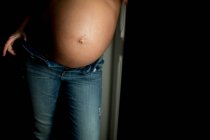 Vientre de mujer embarazada anónima en jeans de pie cerca de la puerta abierta en casa - foto de stock