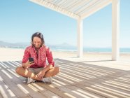 Uomo barbuto smartphone di navigazione mentre seduto in gazebo sulla spiaggia di sabbia vicino al mare nella giornata di sole — Foto stock