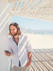 Handsome bearded man using smartphone while leaning on pillar of white gazebo on sandy beach — Stock Photo