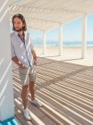 Красивий бородатий чоловік дивиться геть, спираючись на стовп білої альтанки на піщаному пляжі — стокове фото