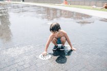 Little boy in swimsuits bending down near jet of water splashing out of fountain on street — Stock Photo