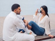 Vue latérale du jeune couple assis et s'amusant à prendre des photos sur banc en bois sur bord de mer sablonneux — Photo de stock