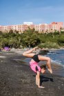Vista lateral de una joven mujer flexible de pie en posición de yoga en la orilla del mar en un día soleado - foto de stock
