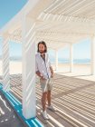 Красивий бородатий чоловік дивиться геть, спираючись на стовп білої альтанки на піщаному пляжі — стокове фото