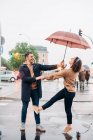 Весёлый молодой человек и женщина с зонтиком обнимаются и смотрят друг на друга, стоя на улице в дождливый день — стоковое фото