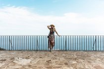 Жінка в літній сукні і солом'яному капелюсі, що стоїть на паркані на кам'яній терасі з морським пейзажем на фоні — стокове фото