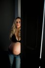 Attractive pregnant woman in bra looking at camera while standing near open door at home — Stock Photo