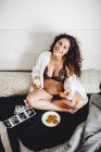 Беременная женщина в нижнем белье сидит, скрестив ноги с помощью ультразвукового сканирования во время перекуса в спальне — стоковое фото