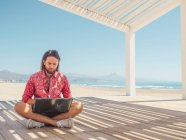 Uomo barbuto che naviga computer portatile mentre seduto in gazebo sulla spiaggia di sabbia vicino al mare nella giornata di sole — Foto stock
