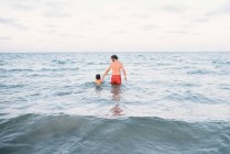 Обратный вид мужчины с мальчиком, держащимся за руки и идущим в воду, плавающим вместе — стоковое фото