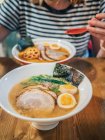 Cropped view of woman sitting at wooden table and enjoying bowl of delicious ramen topped with egg and sliced pork — Stock Photo