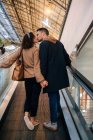 Back view of young man and woman holding hands and kissing while standing on moving walkway during date in brightly illuminated mall — Stock Photo