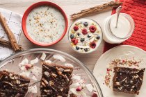 Сверху аппетитный ароматный йогурт с малиновым овсянкой в стекле и красными чашами, украшенными корицей и нарезанным коричневым тортом на деревянном фоне — стоковое фото