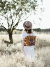Woman in hat with trendy backpack walking among wild field in daylight — Stock Photo