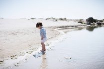 Vista lateral do bebê bonito de pé com os pés descalços na areia e olhando para o mar calmo brilhante na bela praia no dia ensolarado — Fotografia de Stock