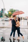 Vista lateral de alegre joven hombre y mujer con paraguas abrazándose y mirándose mientras están de pie en la calle - foto de stock