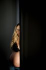 Donna incinta in reggiseno guardando la fotocamera mentre in piedi vicino alla porta aperta nella stanza buia — Foto stock
