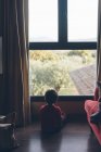 Vista posteriore del piccolo ragazzo sognante seduto sul pavimento in soggiorno e guardando lontano in finestra — Foto stock