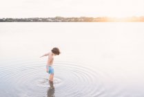 Visão traseira do menino sonhador andando criando ondulações em águas rasas da praia contra a luz do pôr do sol — Fotografia de Stock