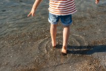 Crop back view of little boy playing in shallow water stepping forward in sunshine — Stock Photo