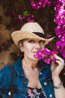 Femme portant un chapeau regardant la caméra tout en se tenant près du mur avec des arbustes de fleurs roses — Photo de stock
