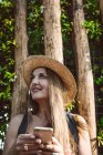 Femme adulte souriante en chapeau de paille tenant smartphone et regardant loin contre les arbres verts — Photo de stock