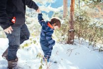 Joven con ropa de abrigo oscuro cogido de la mano con un niño en un mono a rayas y caminando en la nieve en un día soleado - foto de stock