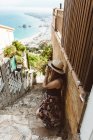 Woman in summer outfit taking photo while standing on street stone stairs with sea coast on background — Stock Photo